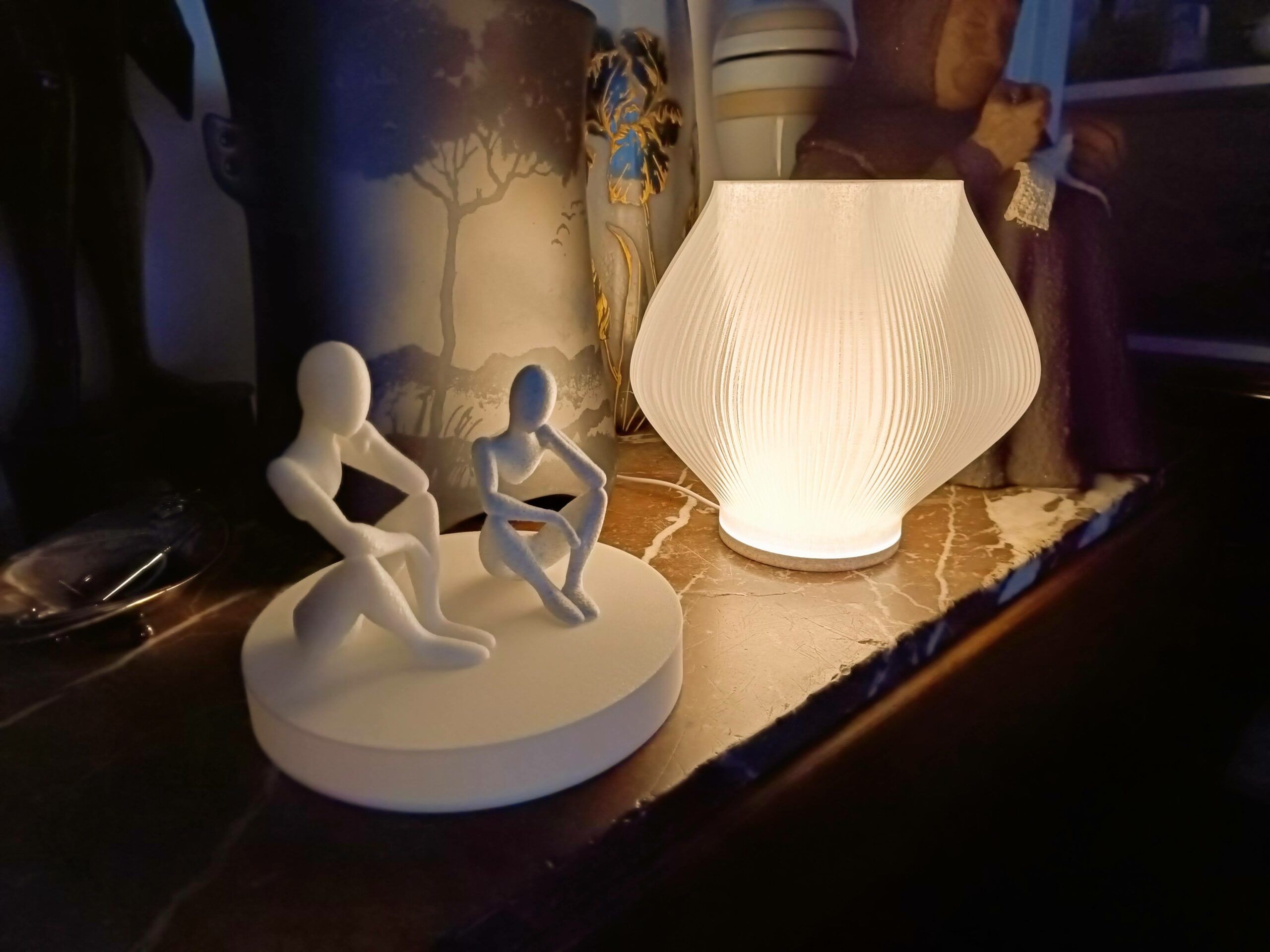 A glowing ribbed white lamp and a sculpture of two seated figures on a round base, both 3D printed, on a marble mantelpiece