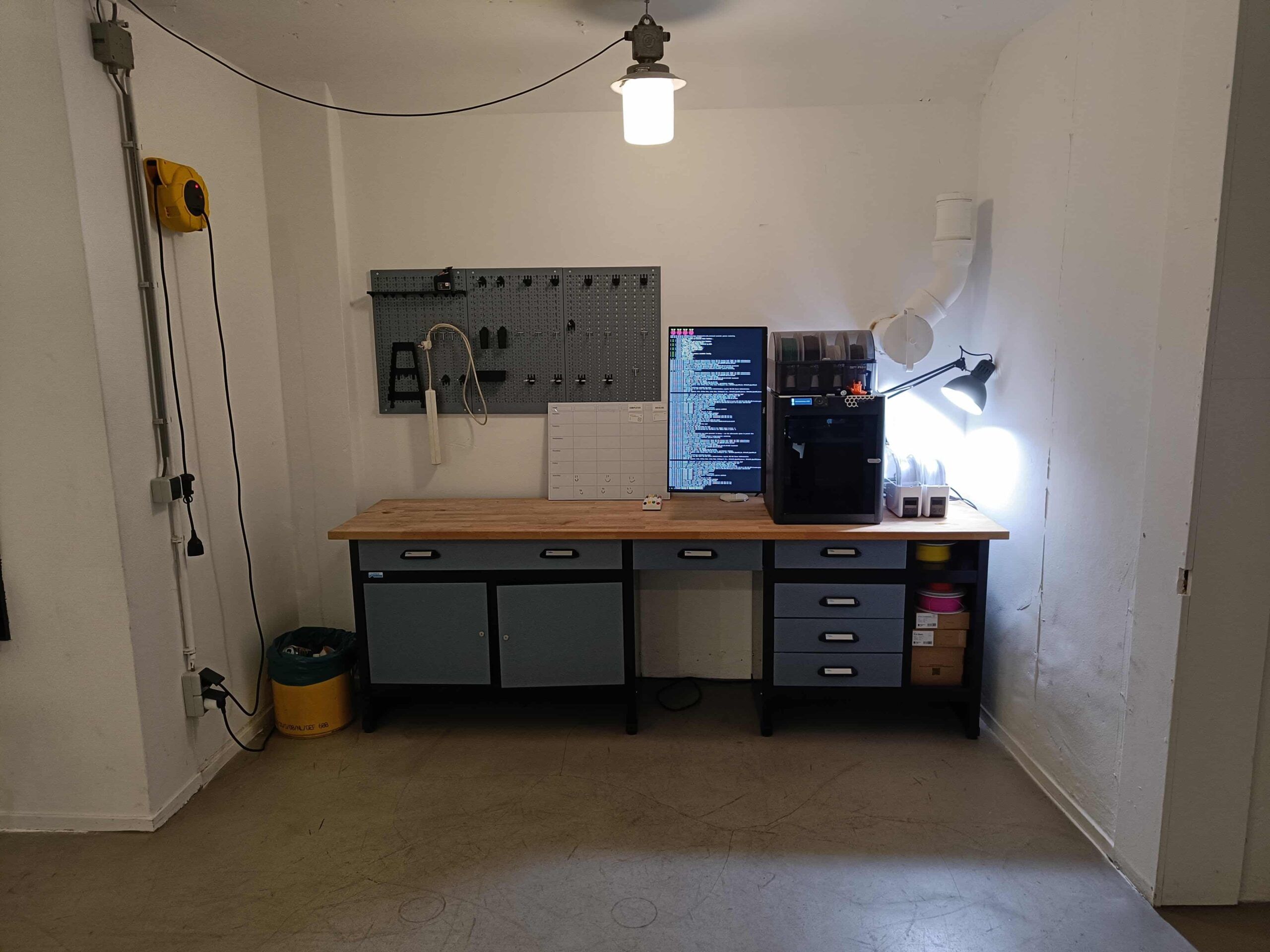 A workbench with a Bambu Lab printer, a material system on top, spools of filament and a pegboard